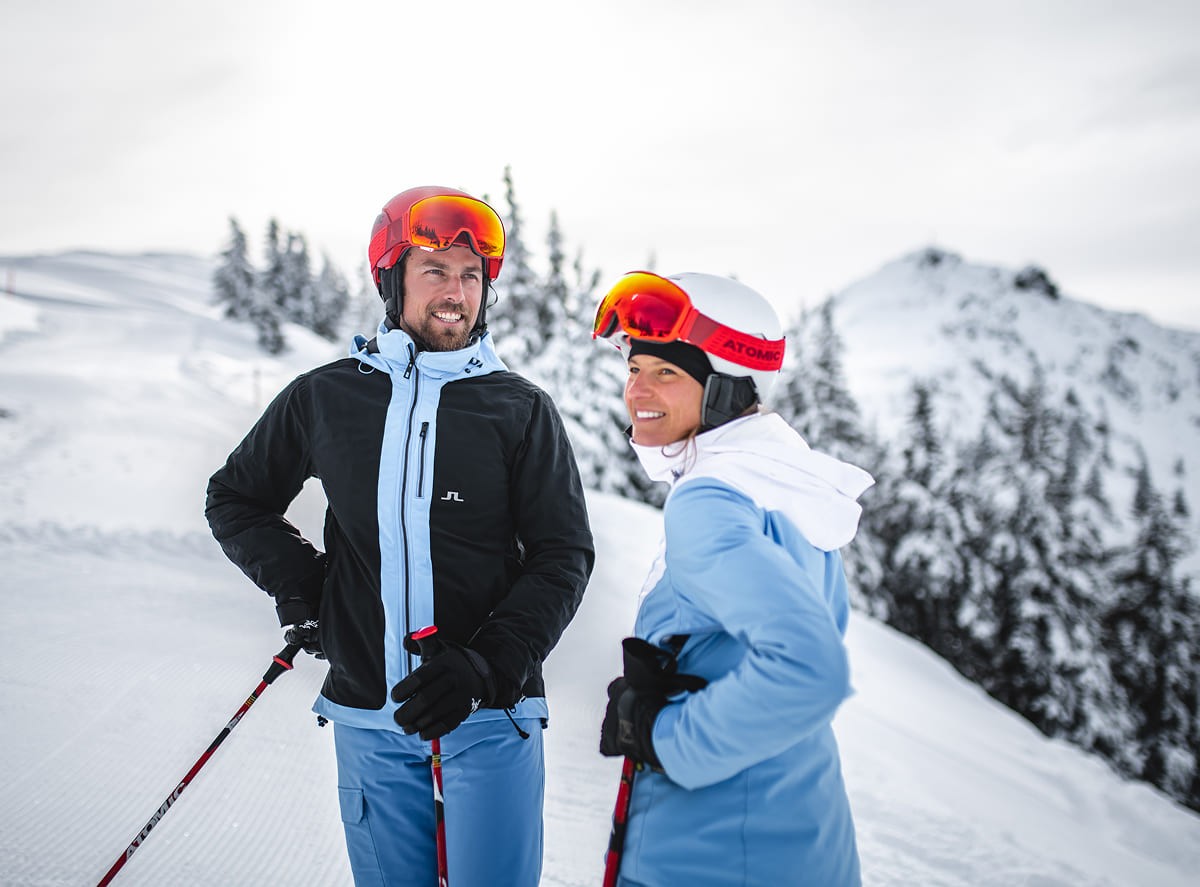 Skifahrer-Paar macht Pause auf der Skipiste mit verschneiter Berglandschaft im Hintergrund © Wagrain-Kleinarl